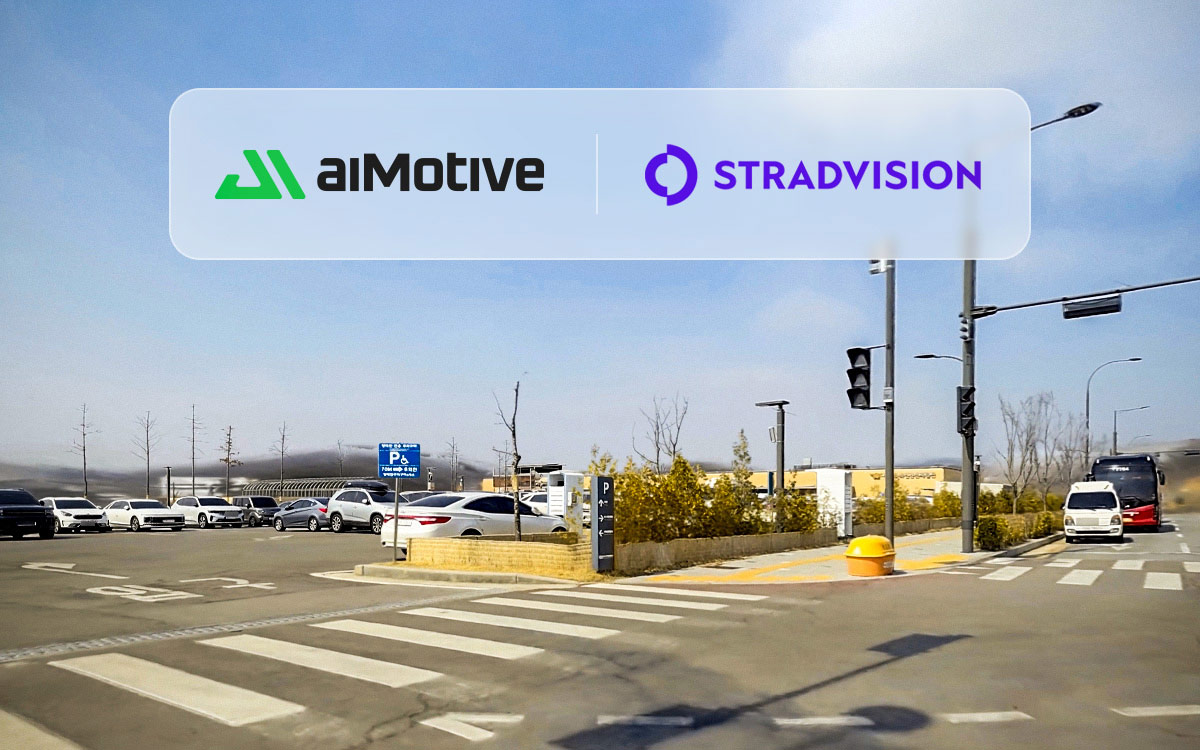 A wide outdoor parking lot at a road intersection on a clear day, with multiple parked cars on the left and a bus approaching from the right near a crosswalk. An overlaid banner displays two company logos: “aiMotive” on the left in green and black, and “STRADVISION” on the right in purple, separated by a vertical line.